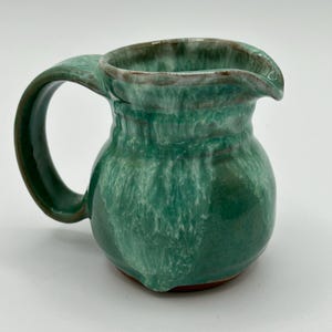 May include: A small green ceramic creamer with a white glaze and a brown rim. The creamer has a rounded body and a handle.