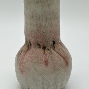 May include: A small, white ceramic vase with a bulbous base and a textured surface. The vase has a pink glaze that drips down the sides.