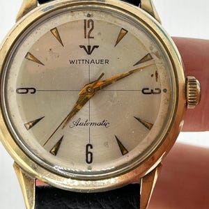 May include: A gold-tone Wittnauer automatic wristwatch with a white face and black leather strap. The watch face has black Roman numeral hour markers and a black second hand.