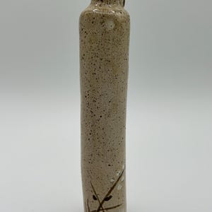 May include: A small, light brown ceramic vase with a textured surface and a handle. The vase has a simple design with brown and white painted flowers and leaves.