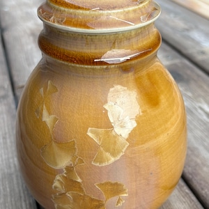 May include: A lidded ceramic jar in warm, earthy tones. The jar has a rounded form and a lid with a stepped design. The surface is decorated with a crackle glaze and crystalline patterns, creating a unique visual texture and appeal.