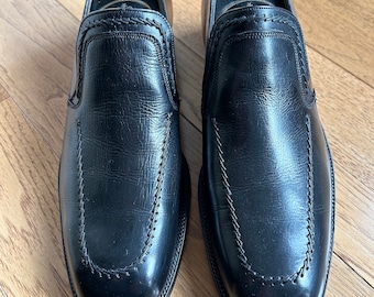 Puede incluir: Un par de zapatos negros de cuero sin cordones con punta cuadrada y un detalle cosido alrededor de la abertura.