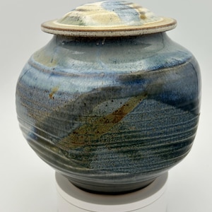 May include: A handcrafted ceramic urn with a lid. The urn has a rounded body with a blue, brown, and tan colour scheme. The lid has a flat top with a similar colour palette. The urn is sitting on a white base.