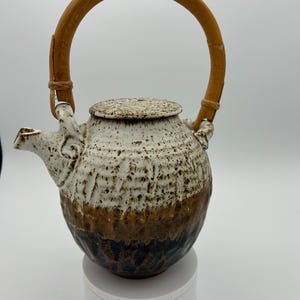 May include: A ceramic teapot with a speckled white, brown, and dark blue glaze. It has a bamboo handle and a matching lid. The teapot is designed for brewing tea.