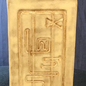 May include: A cream-coloured ceramic vase with a rectangular shape and an embossed design. The design features a series of geometric shapes and lines.