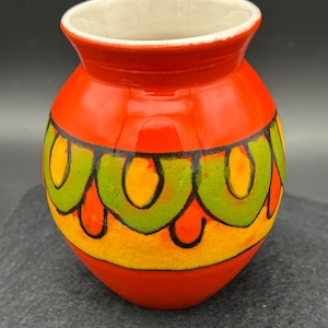 May include: A small, round, ceramic vase with a vibrant orange base and a white interior. The vase features a decorative band of green, yellow, and black curved patterns. The vase is a decorative item.