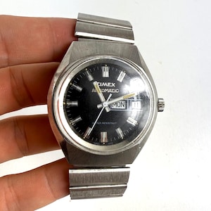 May include: A vintage Timex Automatic watch with a black face and silver-tone metal band. The watch face displays the day and date, with the word "Timex" and "Automatic" visible. The watch band is approximately 0.75 inches wide.