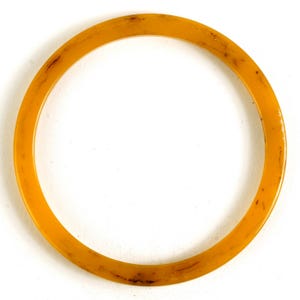 May include: A round, amber-colored bangle bracelet. The bracelet has a smooth, polished surface with subtle variations in color and small dark spots. The bangle is a simple, classic design, suitable for everyday wear.
