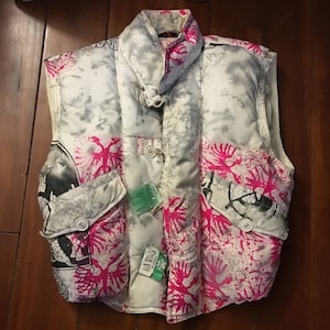 May include: A white, gray, and pink puffer vest with a unique pattern. The vest features a mix of abstract gray shapes, pink coral-like designs, and black graphic elements. It has two front pockets and a high collar. A green tag is attached.