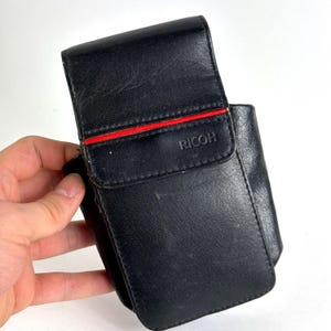 May include: Black leather camera case with a red stripe and the brand name "RICOH" printed on the front. The case has a flap closure and appears to be vintage. The case is rectangular and has a pocket on the side.