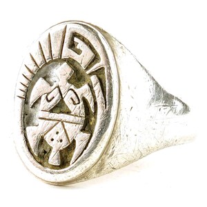 May include: A silver ring with an oval face featuring an engraved design. The design includes geometric shapes and a central figure. The ring has a textured, worn appearance, suggesting it is vintage or antique.