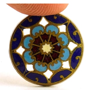 May include: A round, ornate, enameled pendant with a gold-tone metal frame. The design features a central starburst in cream, surrounded by burgundy, light blue, and dark blue floral and geometric shapes. The pendant is held by a finger.
