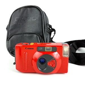 May include: A red Canon film camera with a black lens and flash, accompanied by a black Casio camera case and a black camera strap. The camera has the word "Canon" printed on the front.