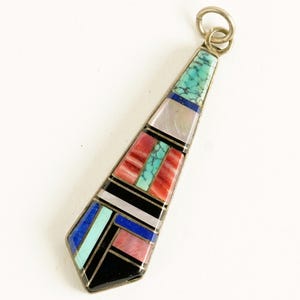 May include: A silver pendant with a geometric design. The pendant features inlaid turquoise, red, black, white, and pink stones. The pendant has a triangular shape with a silver loop at the top for hanging.
