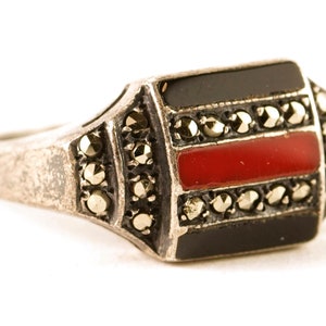 May include: A silver ring with a rectangular black and red stone set in the center. The stone is surrounded by a border of small, sparkling stones.