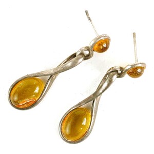 May include: A pair of silver-toned drop earrings with amber-colored, oval-shaped stones. The earrings have a twisted metal design and a post-and-clutch closure. The amber stones are a warm, translucent color.
