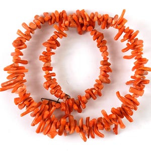 May include: A strand of orange coral beads, strung together to form a necklace. The beads are irregular in shape and size, with some being long and thin, while others are shorter and thicker.