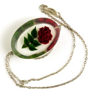 May include: Silver-toned necklace featuring an oval pendant with a red rose and green leaves encased in clear resin. The pendant has a red and green border. The chain is a delicate link style, suitable for everyday wear.