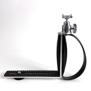 May include: A black and silver camera bracket with a curved handle and a mounting plate. The bracket has a silver top with the word "Accura" printed on it. The mounting plate has a black, textured surface.