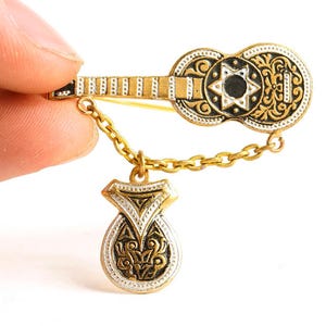 May include: A gold-toned guitar-shaped brooch with black and white accents, connected by a chain to a pendant. The brooch features a star design and ornate detailing. The pendant has a decorative pattern.