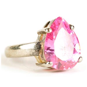 May include: A silver ring featuring a large, pear-shaped pink gemstone. The gemstone is faceted, catching the light and displaying a vibrant color. The ring has a simple band and four prongs securing the stone.