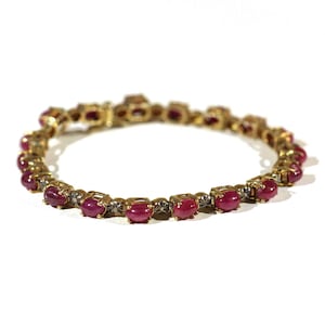 May include: A gold bracelet with a row of oval-shaped ruby stones set in a gold frame. The bracelet is on a white background.