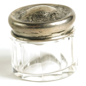 May include: A clear, faceted glass jar with a decorative silver-toned lid. The lid features an embossed design. The jar is likely for storing small items or cosmetics. The jar is on a white background.