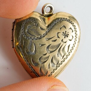 May include: A vintage heart-shaped locket with an ornate floral design. The gold-toned locket is held between two fingers, showcasing the intricate engraved details. The locket has a small loop at the top for a chain.