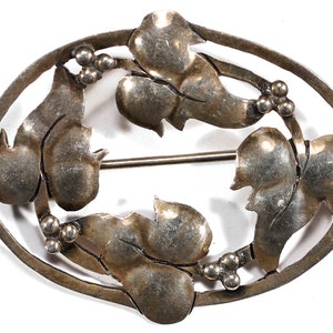 May include: A silver brooch with a design of stylized leaves and berries. The brooch is oval shaped and has a pin on the back.