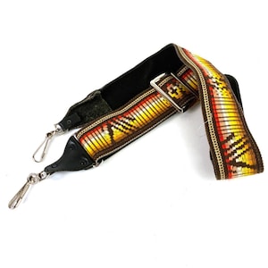 May include: A brown guitar strap with a colorful geometric pattern in red, yellow, and white. The strap has a black leather adjustable section and two silver metal clasps.