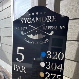 May include: Black metal golf course sign with white text that reads "SYCAMORE", "EST. 1973", "RAVENA, NY", and "COUNTRY CLUB". The sign also has a golf club graphic and a scorecard with the numbers 5, 320, 304, 275, 275, and 4.