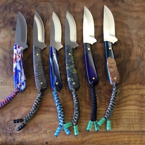 May include: Six custom knives with various handle designs and stainless steel blades. Handles feature a range of colors and patterns, including blue, gray, and wood grain. Each knife has a braided lanyard with decorative beads.