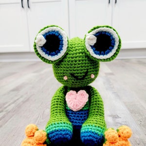 Könnte beinhalten: Ein grüner Häkel-Frosch mit blauen Augen, einem rosa Herz auf der Brust und orangefarbenen Füßen. Der Frosch sitzt auf einem Holzboden. Der Text "YarnByCoco" ist im Bild sichtbar.