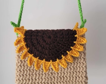 Op de afbeelding: Een gehaakte crossbody tas in de vorm van een zonnebloem met een bruin centrum en gele bloemblaadjes. De tas heeft een groene band en de tekst "YarnByCoco" is zichtbaar aan de onderkant.