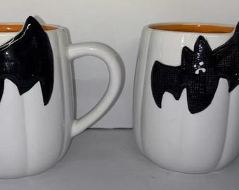 May include: Two white ceramic mugs with orange rims and black bat silhouettes. The mugs are shaped like pumpkins.
