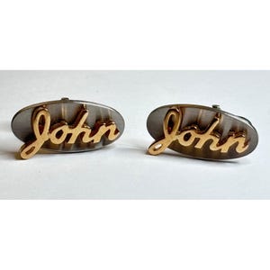 May include: A pair of oval-shaped cufflinks featuring a brushed silver-tone metal base with the name John written in a raised, cursive gold-tone script. The design is identical on both pieces, showcasing a metallic contrast between the silver background and the gold lettering.