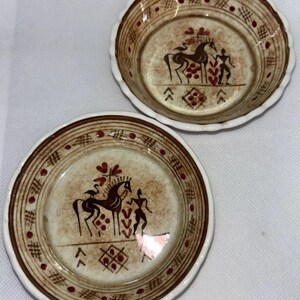 May include: Two small ceramic bowls with a brown and white design. The bowls feature a stylized horse and human figure in red, brown, and white. The bowls have a scalloped edge.