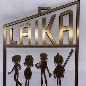 Puede incluir: Un recorte de metal color bronce con la palabra "LAIKA" encima de cuatro figuras estilizadas. Las figuras parecen ser músicos, uno de ellos sosteniendo una guitarra. El recorte tiene forma rectangular con un pequeño lazo en la parte superior.