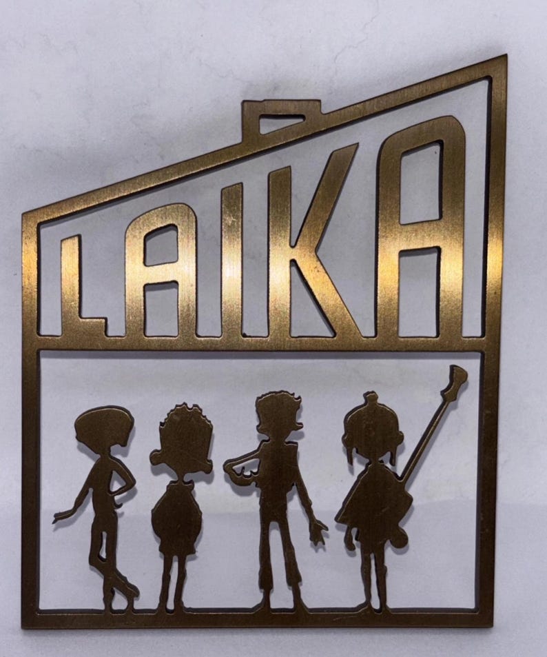 May include: A bronze-coloured metal cutout featuring the word "LAIKA" above four stylised figures. The figures appear to be musicians, with one holding a guitar. The cutout has a rectangular shape with a small loop at the top.