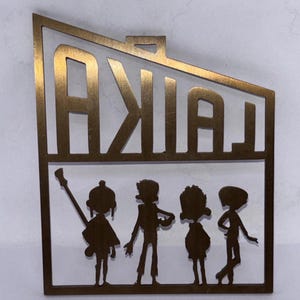 May include: A gold-coloured metal cutout featuring the word "LAIKA" above four stylised figures. The figures appear to be a band, with one holding a guitar. The cutout has a rectangular shape.