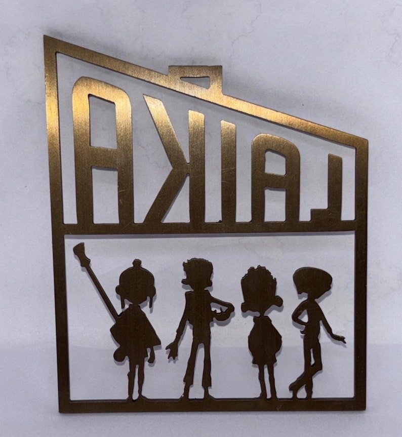 May include: A gold-coloured metal cutout featuring the word "LAIKA" above four stylised figures. The figures appear to be a band, with one holding a guitar. The cutout has a rectangular shape.