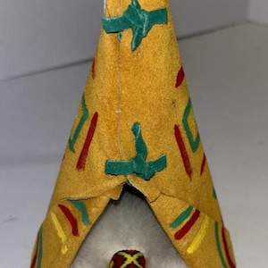 May include: A small, handmade Native American teepee made of suede with red, green, and yellow geometric patterns. The teepee has a white fur lining and a small, brown figure inside wearing a red and yellow headdress.