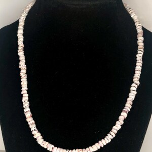 May include: A necklace composed of small, cylindrical white beads featuring irregular brown and tan speckled patterns. The beads are strung together in a single continuous strand, creating a uniform circular shape that rests evenly around the neck.