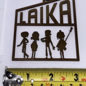 May include: A brown metal cutout with the word "LAIKA" and four stylized figures. The cutout is approximately 5 cm wide. The figures appear to be playing instruments. The cutout is on a white background.