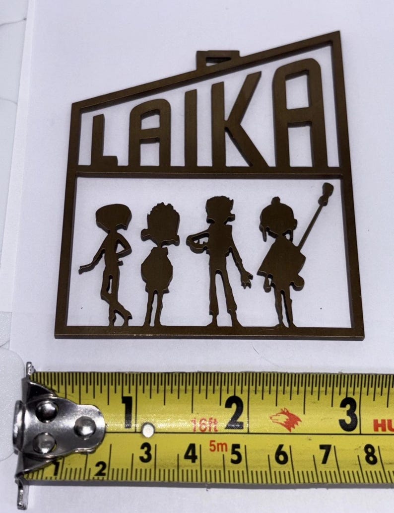 May include: A brown metal cutout with the word "LAIKA" and four stylized figures. The cutout is approximately 5 cm wide. The figures appear to be playing instruments. The cutout is on a white background.