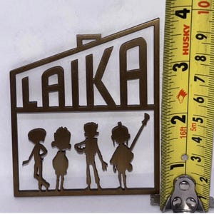 May include: A bronze metal cutout with the word "LAIKA" and four figures. The figures appear to be musicians. The item is approximately 10 cm in height, as shown by the measuring tape.