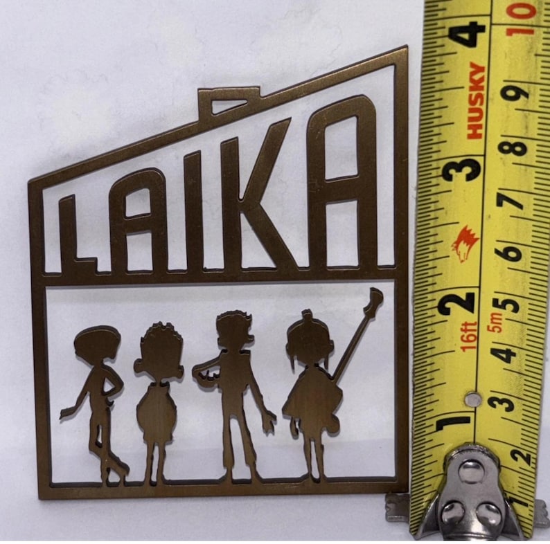 May include: A bronze metal cutout with the word "LAIKA" and four figures. The figures appear to be musicians. The item is approximately 10 cm in height, as shown by the measuring tape.