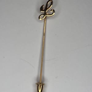 May include: A gold-toned metal stick pin featuring a cursive letter L at the top. The pin has a long, slender straight shaft and a small, rounded, bell-shaped cap at the bottom end. The design is simple, metallic, and polished in appearance.