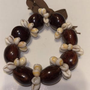 Puede incluir: Un lei circular con cuentas pulidas de color marrón oscuro y pequeñas conchas blancas. Una cinta marrón está atada en la parte superior. Este lei hawaiano tradicional se usa a menudo en celebraciones.