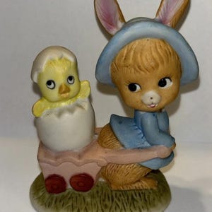 May include: A ceramic Easter figurine featuring a bunny wearing a blue bonnet and dress, pulling a wagon with a yellow chick hatching from an egg. The figurine is set on a green grassy base.